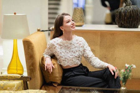 Lächelnde und hellhaarige Frau in Spitzentop und Hose sitzt auf dem Sofa in der Berliner Hotellobby