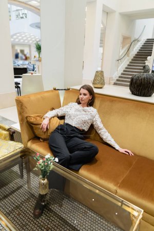 Entspannte blonde junge Frau in Spitzentop und Hose sitzt auf Sofa in Berliner Hotellobby