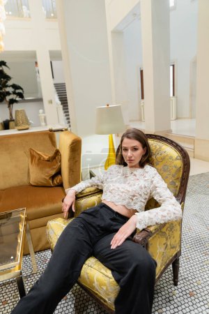 mujer joven y elegante en top de encaje y pantalones mirando a la cámara mientras está sentado en el sillón 