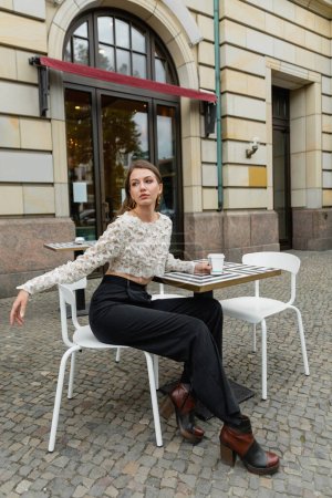 young woman in lace top and pants looking away while holding coffee and sitting near outdoor cafe