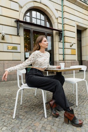 Mujer de moda con top de encaje y pantalones mirando hacia otro lado mientras sostiene el café y se sienta al aire libre