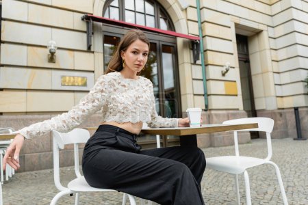 mujer joven en top de encaje y pantalones sosteniendo café y sentado a la mesa de café al aire libre en Berlín 