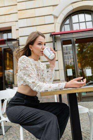 Foto de Mujer joven bebiendo café para ir mientras pasa tiempo en la mesa de café al aire libre - Imagen libre de derechos