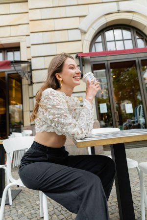 Cheerful young woman in lace top and high waist pants holding coffee to in Berlin, Germany 