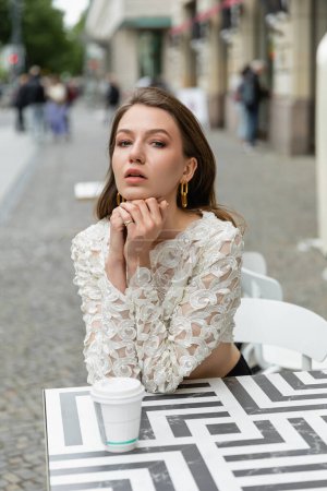 Portrait of fashionable and pretty young woman in white lace top sitting near coffee to go on table