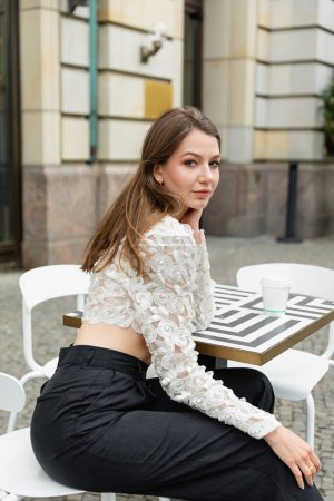 Young and fair haired woman in lace top and pants looking at camera while sitting near coffee to go