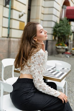 Selbstbewusste und modische junge Frau in Spitzentop und Hose, die neben Kaffee sitzt, um auf den Tisch zu gehen