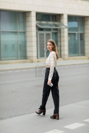Volle Länge der modischen jungen Frau in Spitzentop und Hose, die in die Kamera schaut und Kaffee to go in der Hand hält