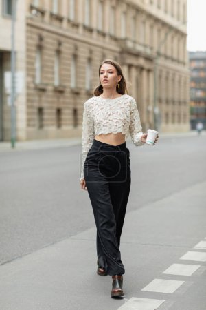 mujer con estilo en la parte superior de encaje y pantalones de cintura alta sosteniendo café para ir a la calle en Berlín, Alemania 