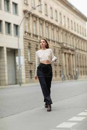 young woman in lace top and high waist pants holding coffee to go on street in Berlin, Germany 