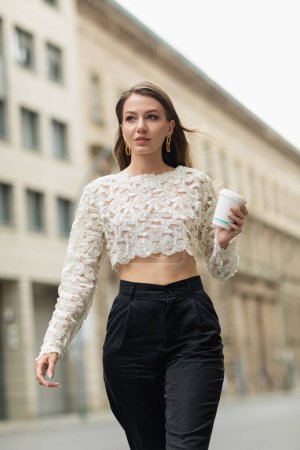 mujer confiada en encaje superior y pantalones de cintura alta sosteniendo café para salir a la calle en Berlín, Alemania 