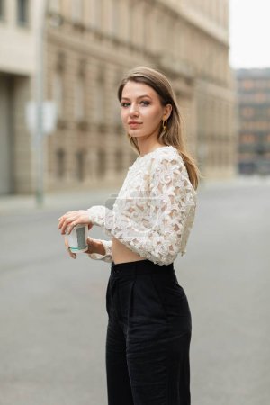 Retrato de una joven mujer de pelo justo en pantalones de cintura alta y top de encaje sosteniendo café para ir en Berlín