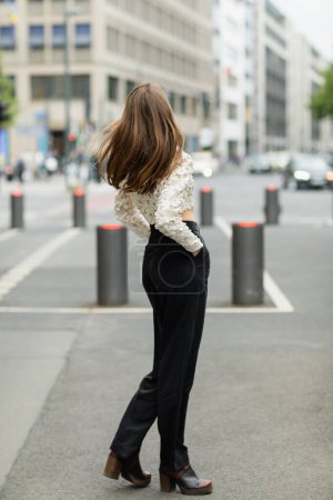 Back view of young fair haired woman in lace top and high waist pants standing on street in Berlin