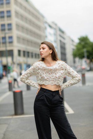 trendy young woman posing in lace top and high waist pants standing on street in Berlin, Germany