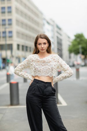confident young woman posing in lace top and high waist pants standing on street in Berlin, Germany
