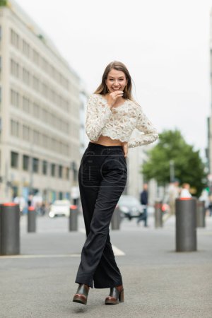 happy young woman posing in lace top and high waist pants standing on street in Berlin
