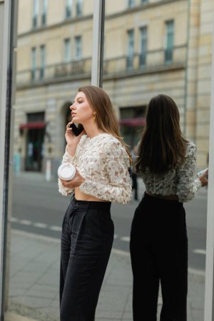 Joven mujer de moda sosteniendo café para ir y hablar en el teléfono inteligente en Berlín, Alemania 