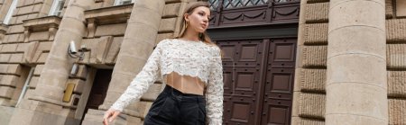 young woman in lace top and high waist pants looking away on urban street in Berlin, banner