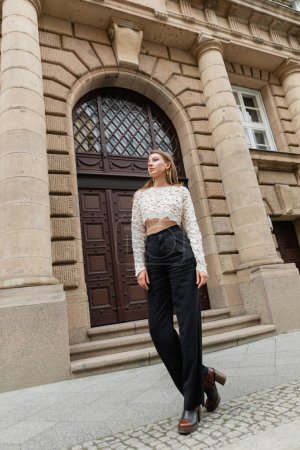 confident stylish woman in lace top and high waist pants looking away on urban street in Berlin