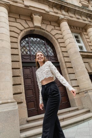 mujer joven optimista en top de encaje y pantalones de cintura alta mirando a la cámara en la calle urbana de Berlín