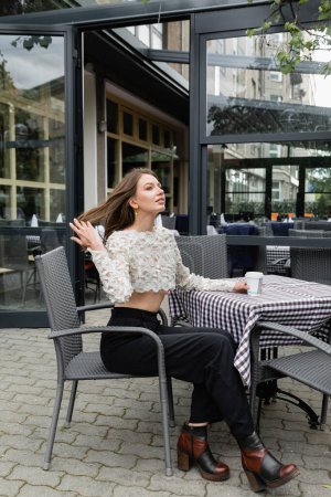Foto de Mujer con estilo tocando el cabello y sosteniendo el café para ir mientras está sentado en la terraza de la cafetería al aire libre - Imagen libre de derechos