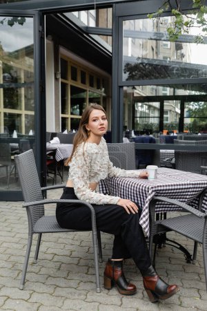 young woman sitting with paper cup on terrace of outdoor cafe and urban street in Berlin