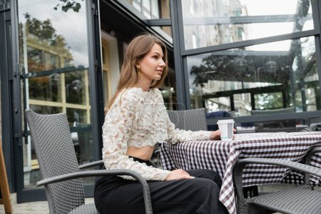stylish young woman sitting with paper cup on terrace of outdoor cafe and urban street in Berlin