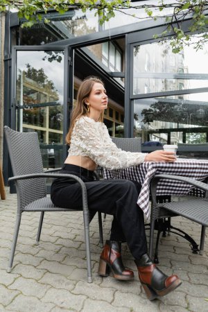 Trendy young woman holding coffee to go while sitting at table on terrace near cafe in Berlin