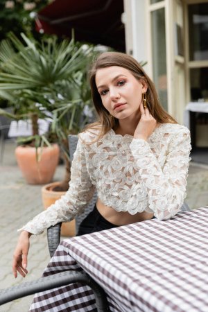 Portrait of young woman in stylish lace top touching neck while sitting near table on terrace