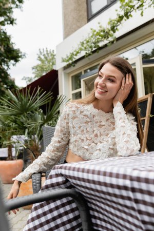 Positive young fair haired woman in stylish lace top sitting near table on cafe terrace
