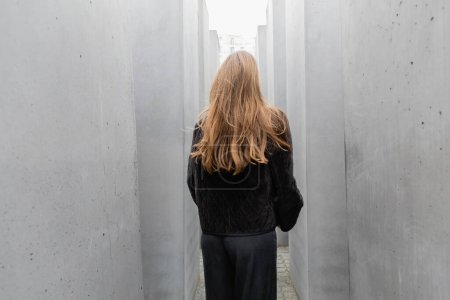 Vista posterior de la mujer en chaqueta negra caminando entre piedras de Memorial to Murdered Jews of Europe