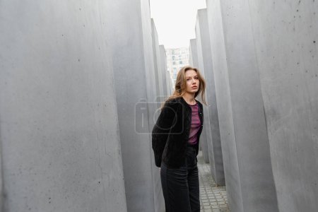 Eine blonde junge Frau in Jacke steht zwischen dem Denkmal für die ermordeten Juden Europas in Berlin