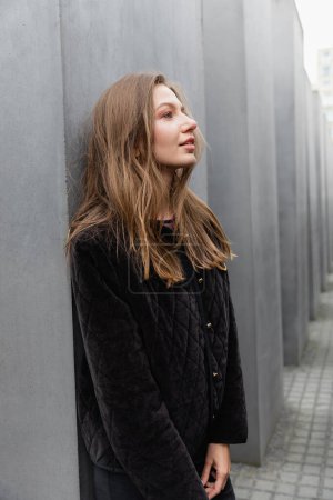fair haired relaxed  woman in jacket standing between Memorial to Murdered Jews of Europe in Berlin