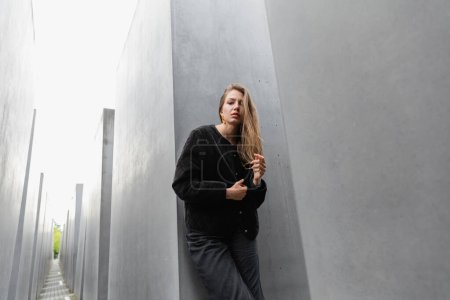 trendy young woman in jacket standing between Memorial to Murdered Jews of Europe in Berlin