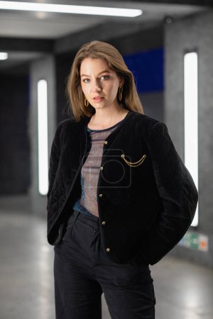 stylische junge Frau in schwarzer Jacke posiert in modernem Gebäude mit Leuchtstoffröhren in Berlin