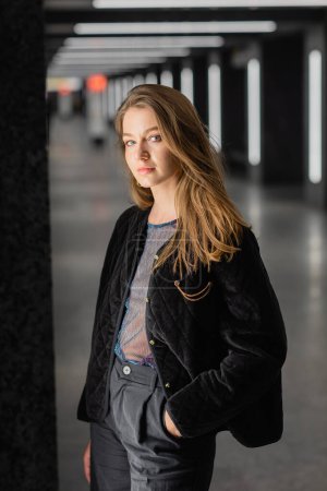 atractiva joven en chaqueta negra posando en edificio moderno con lámparas fluorescentes en Berlín