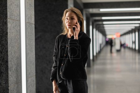 Femme élégante en veste noire parlant sur smartphone tout en se tenant à l'intérieur du bâtiment moderne