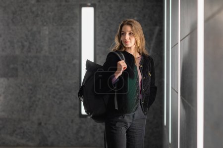 Mujer de moda en chaqueta negra sosteniendo la mochila y mirando hacia otro lado en el edificio moderno 