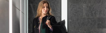 Trendy young fair haired woman in black jacket holding backpack and looking away, banner 