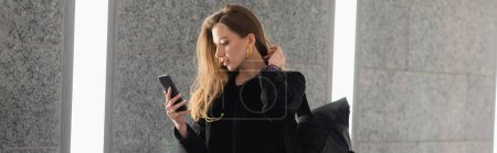 Femme tendance en veste avec sac à dos en utilisant un téléphone portable tout en se tenant près du mur en béton gris, bannière
