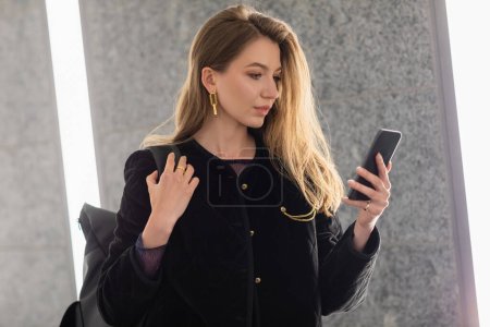 young woman in black jacket holding backpack and using smartphone near lighting of fluorescent lamps