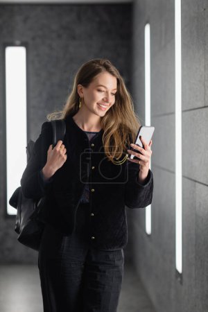 mujer feliz en la chaqueta que sostiene la mochila y el uso de teléfono inteligente cerca de la iluminación de lámparas fluorescentes 