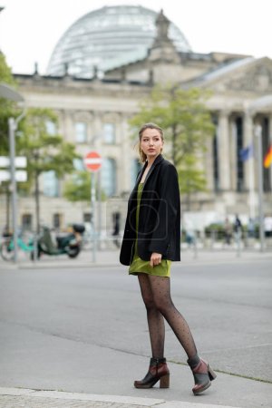 Full length of stylish woman in green silk dress, boots and jacket looking away outdoors