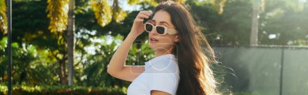 Foto de Mujer morena cautivadora y joven con el pelo largo de pie en camiseta blanca y gafas de sol de moda cerca de palmeras borrosas y verdes en Miami, día soleado, viajes, vacaciones, pancarta - Imagen libre de derechos