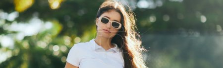 charming and young woman with brunette long hair standing in white polo shirt and trendy sunglasses near blurred and green palm trees in Miami, sunny day, travel, vacation, banner, sun-kissed 
