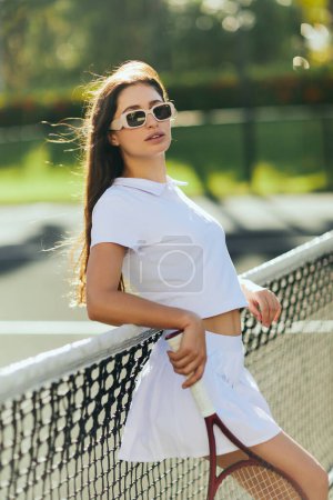 Téléchargez les photos : Court de tennis à Miami, jeune femme sportive aux cheveux longs bruns debout en tenue blanche et lunettes de soleil tout en tenant la raquette près du filet de tennis, fond flou, ville emblématique, regardant la caméra - en image libre de droit