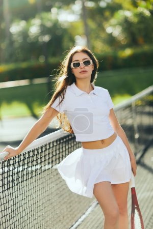 court de tennis à Miami, belle joueuse aux cheveux bruns debout en tenue blanche et lunettes de soleil tout en tenant la raquette près du filet de tennis, fond flou, ville emblématique, regardant la caméra 