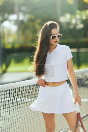 pista de tenis en Miami, mujer joven deportiva con pelo largo morena de pie en traje blanco y gafas de sol mientras sostiene raqueta cerca de la red de tenis, fondo borroso, ciudad icónica, Florida 