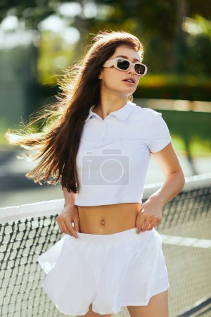 portrait de jeune femme aux cheveux longs bruns debout en tenue blanche et lunettes de soleil près du filet de tennis, fond flou, vent, court de tennis à Miami, ville emblématique, joueuse, Floride 