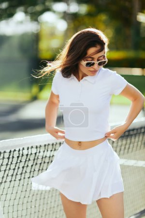 Porträt einer hübschen jungen Frau mit brünetten langen Haaren im weißen Outfit und Sonnenbrille in der Nähe des Tennisnetzes, verschwommener Hintergrund, Wind, Tennisplatz in Miami, ikonische Stadt, Spielerin, Florida 
